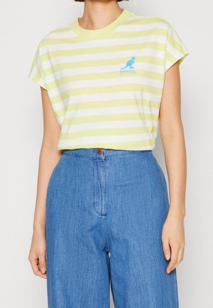 Osoba ubrana w krótki rękaw, żółto-białą pasiastą koszulkę Kangol wpuszczoną do wysokich, niebieskich dżinsów.