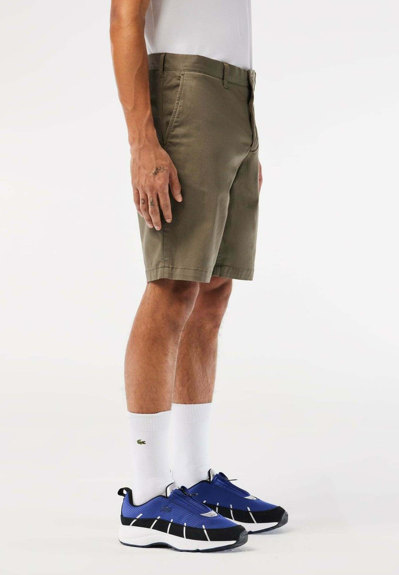 Shorts en khaki avec une taille plate et des poches, associés à des chaussures de sport bleues et des chaussettes blanches. Tissu lisse, design structuré.