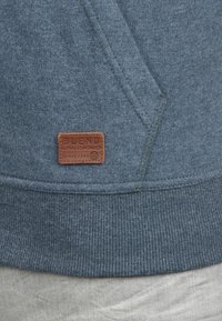Sudadera de forro polar azul con dobladillo acanalado y una etiqueta de cuero marrón con "BLEND" en relieve. Tejido texturizado con un acabado suave.
