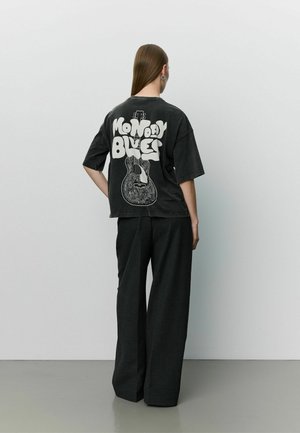 Personne aux cheveux longs portant un pantalon noir à jambes larges et un t-shirt noir avec "Monday Blues" et un motif de guitare dans le dos, debout contre un mur uni.