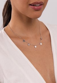 Silberne Halskette mit einem Kettendesign, das fünf kleine, runde Anhänger enthält. Glatte Oberfläche, um den Hals getragen, über einem hellen Oberteil.