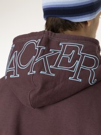 Burgunderfarget hettegenser med blå brodering som danner ordet "HACKER" på baksiden. Har snor i hetta og myk tekstur.