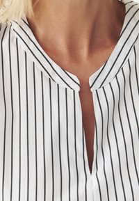 Valkoinen paita, jossa on pystysuoria mustia raitoja, kauluksella ja halkaistulla kaula-aukolla. Pehmeä kangas ja sileä tekstuuri ilman lisäkuvioita.