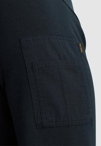 Pantaloni cargo blu navy scuri con una tasca laterale con bordi cuciti e un piccolo etichetta del marchio in giallo senape. Materiale in cotone morbido e testurizzato.