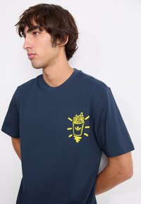 Marine t-shirt lavet af bomuld med et gult grafisk motiv af pomfritter i en kop på venstre bryst med strålende linjer omkring.