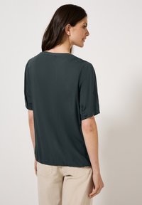 Blusa corta a maniche corte di colore verde scuro con scollatura rotonda. Tessuto liscio, vestibilità leggermente ampia e maniche con bottoni. Design leggero e informale.