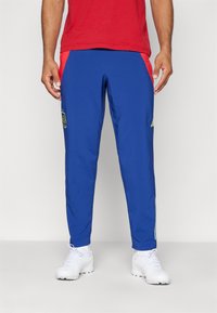 Pantalones deportivos azules con paneles laterales rojos. Presenta un logo bordado en la pierna izquierda y franjas blancas a lo largo de los lados. Hechos de material ligero.