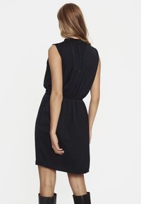 Svart ärmlös klänning med veckad rygg, markerad midja, knälång, slät tyg, med en subtil drapering och nyckelhålsstängning vid halsen.