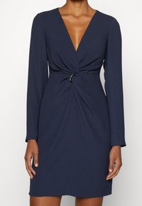 Marinblå klänning med djup V-ringning, långa ärmar, vriden detalj framtill, markerad midja och silverfärgad detalj vid midjan.