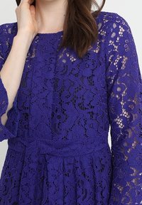 Fialové krajkové šaty s propracovanými květinovými vzory, dlouhými rukávy, kulatým výstřihem a přiléhavým pasem. Jemná, strukturovaná tkanina.