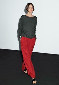 Une femme marche avec confiance dans un pull gris et un pantalon rouge vif, sur un fond minimaliste et un sol noir, mettant en avant un style contemporain.