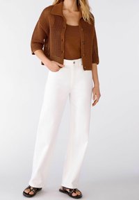 Hnědý pletený krátký kardigan přes hnědý top na ramínka, doplněný vysokými bílé široké džíny a černými sandály.