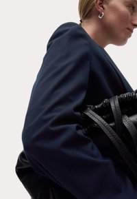 Blazer bleu marine avec épaules structurées, associé à un sac à main en cuir noir avec un dessus resserré et une bandoulière texturée. Boucle d'oreille en argent visible.