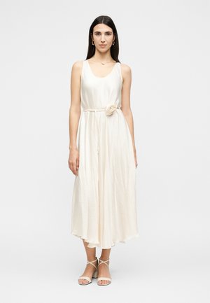 Femme aux longs cheveux foncés portant une robe midi sans manches couleur crème avec une ceinture à la taille et des sandales beiges à talons blocs à brides, faisant face.