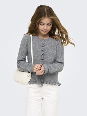 Djevojka u sivoj jakni s gumbima i naborima uz rubove, bijelim hlačama i bijelom torbom preko ramena, gleda prema dolje i drži ruke.