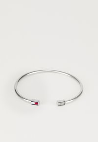 Pulsera de brazo de metal plateado con un diseño abierto, que presenta extremos cuadrados; un extremo tiene un acento rojo y blanco, el otro es liso.