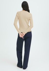Donna con lunghi capelli scuri in piedi con le mani incrociate dietro la schiena, vestita con un maglione a coste beige e pantaloni larghi blu navy.