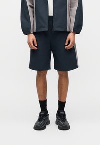 Άνδρας που φοράει μπλε ναυτικό σορτς, μαύρες κάλτσες, μαύρα αθλητικά παπούτσια Diesel και μπλε ναυτικό και γκρι μπουφάν με φερμουάρ, σε απλό ανοιχτό φόντο.