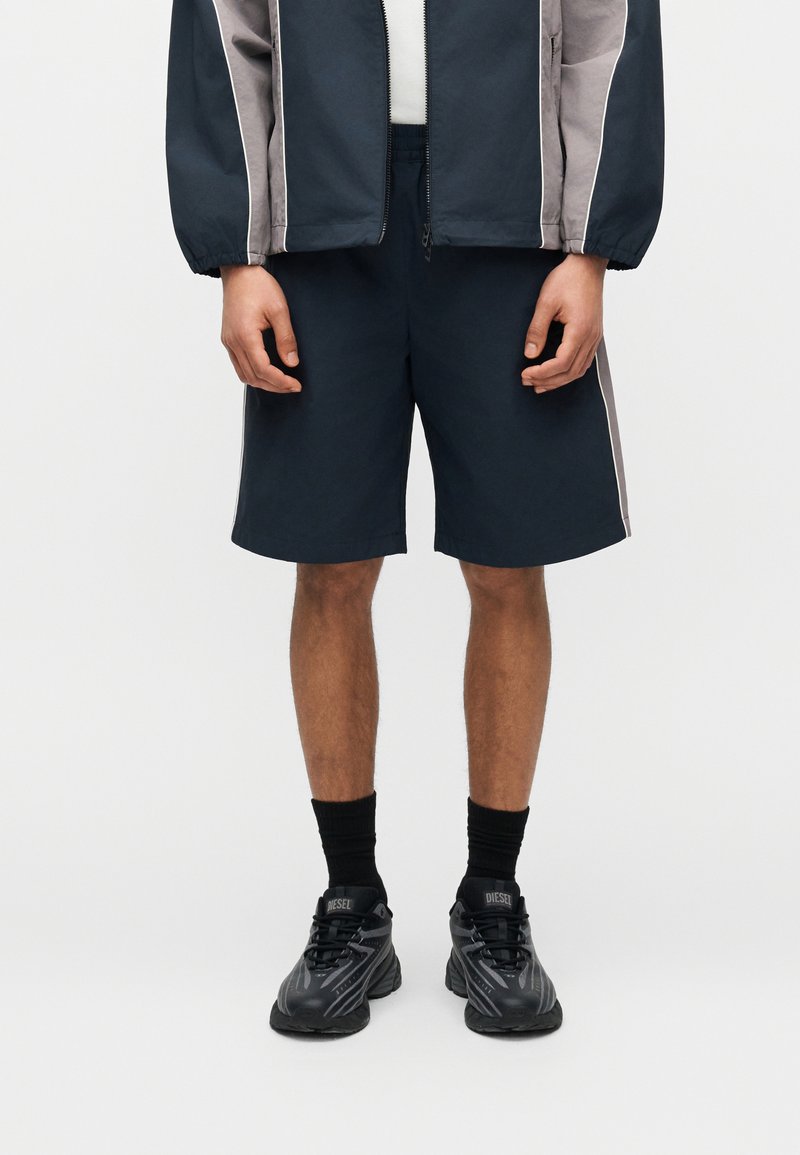 Άνδρας που φοράει μπλε ναυτικό σορτς, μαύρες κάλτσες, μαύρα αθλητικά παπούτσια Diesel και μπλε ναυτικό και γκρι μπουφάν με φερμουάρ, σε απλό ανοιχτό φόντο.