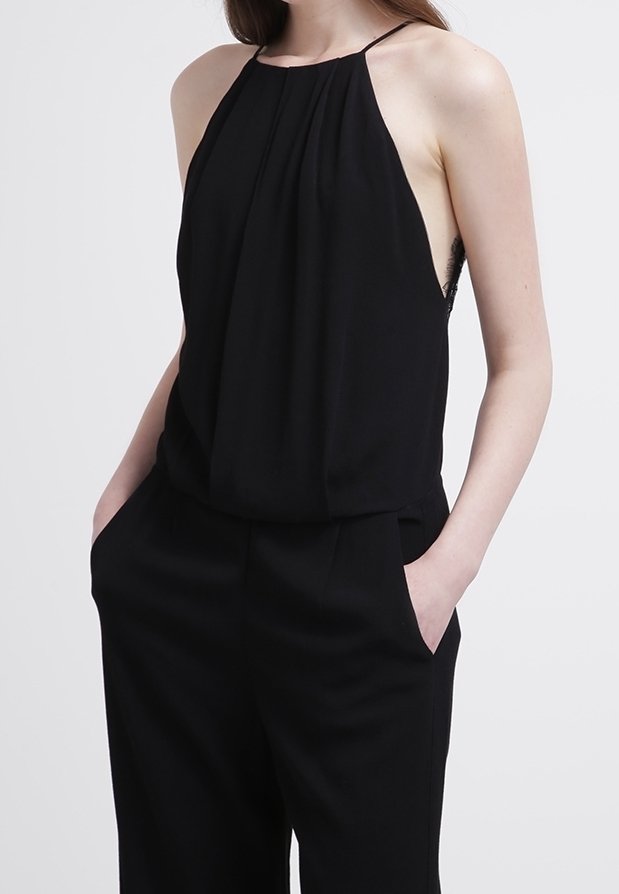 Femme portant un haut noir sans manches à col halter et un pantalon noir large, les mains dans les poches, sur un fond uni.