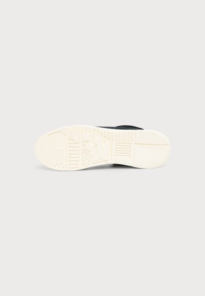 Semelle en caoutchouc blanche d'une chaussure noire vue de dessous avec des motifs de grip texturés sur un fond gris clair.