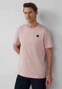 Hellrosa Baumwoll-T-Shirt mit kurzen Ärmeln, Rundhalsauschnitt und einem schwarzen quadratischen Emblem auf der Brust. Getragen mit dunklen Jeans und neutralem Hintergrund.