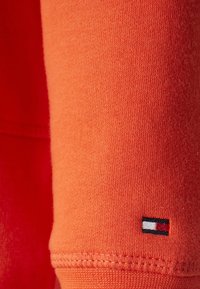 Tela de algodón naranja con textura suave, que presenta un pequeño logo bordado en negro, blanco y rojo en el puño.