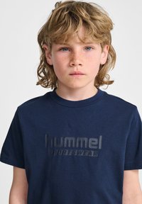 Granatowa bawełniana koszulka z krótkim rękawem, z podniesionym czarnym logo z napisem "hummel SPORTSWEAR". Okrągły dekolt.