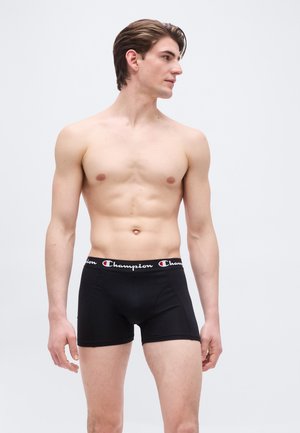 Een shirtloze man die zwarte Champion boxershorts draagt, staat met het gezicht naar links tegen een effen lichte achtergrond.