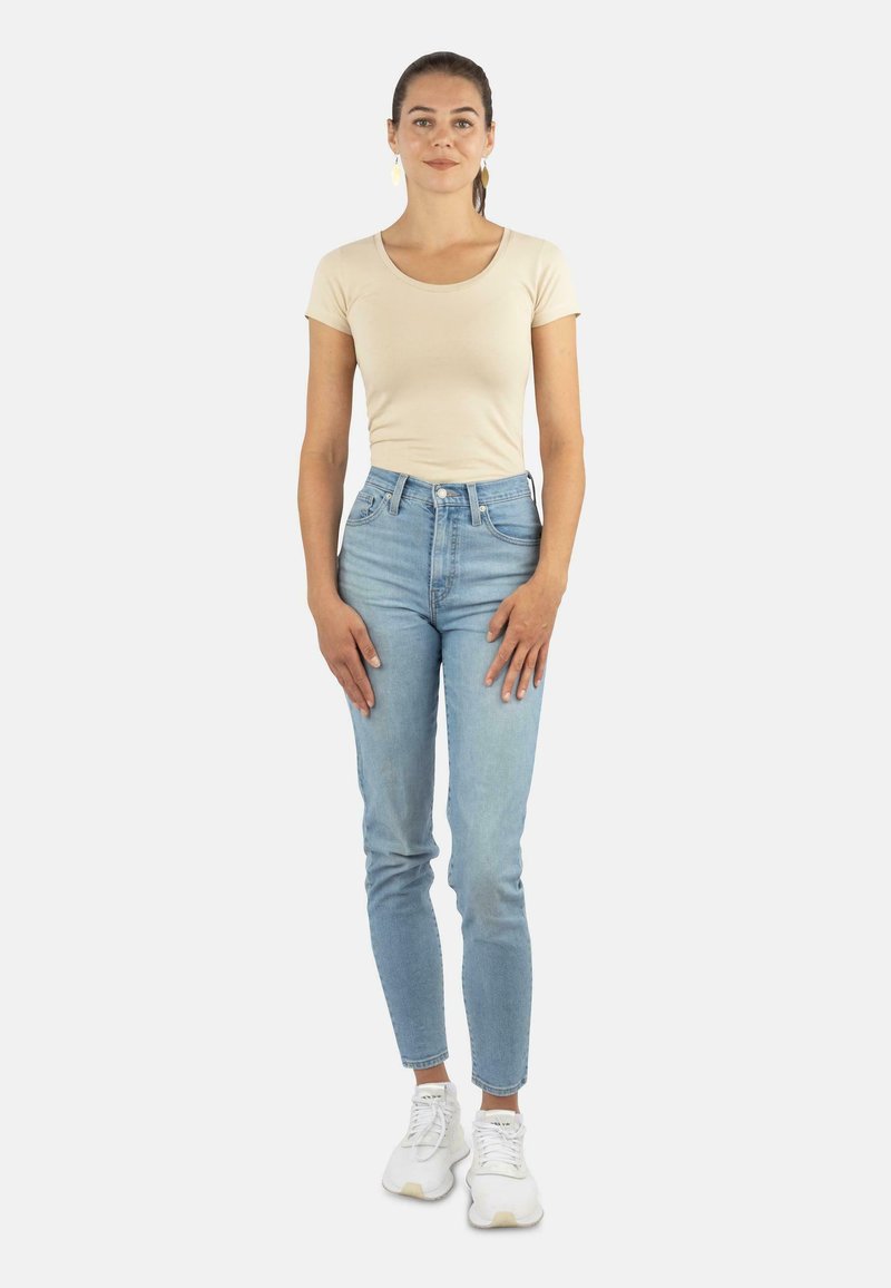 Beige eng anliegendes T-Shirt, hochgeschnittene hellblaue Jeans und weiße Sneakers. Das Modell steht aufrecht mit den Händen an den Seiten vor einem schlichten Hintergrund.