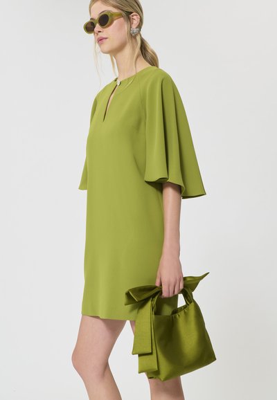 Femme en robe vert olive à manches larges et encolure goutte, tenant un sac à main en tissu assorti et portant des lunettes de soleil vertes.