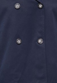 Ύφασμα ναυτικού μπλε με τέσσερα μεγάλα στρογγυλά σκουρόχρωμα κουμπιά, διατεταγμένα σε διπλόστηλη διάταξη, ορατές κουμπότρυπες ραμμένες.