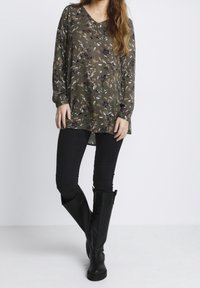 Olijfgroene bloes met lange mouwen, V-hals en een losse pasvorm, gecombineerd met zwarte skinny jeans en kniehoge zwarte laarzen.