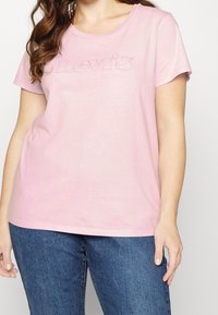 Světle růžové tričko s krátkým rukávem z měkkého materiálu s kulatým výstřihem, na kterém je vyšitý název značky "Levi's" v tmavším odstínu.