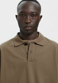 Brūns polo krekls ar teksturētu audumu, tam ir apkakle, divas pogas un neliels plākstera detaļa. Piedurkņu griezums ir brīvs un ikdienišķs.