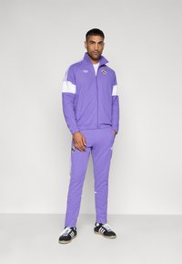 Αθλητική φόρμα σε λιλά χρώμα με λευκές ρίγες, μπουφάν με φερμουάρ και ψηλό γιακά, ταιριαστό παντελόνι και μαύρα αθλητικά παπούτσια με λευκές λεπτομέρειες.