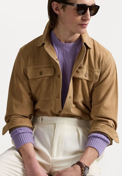 Braunes Cordhemd mit zwei Fronttaschen, übereinander getragen mit einem lavendelfarbenen Strickpullover, kombiniert mit cremefarbenen Hosen und dunklen Sonnenbrillen.