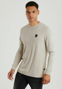 Langärmliges beige Hemd aus weichem Baumwollstoff, mit Rundhalsausschnitt, schmalen Bündchen und einem kleinen schwarzen Logo-Patch auf der Brust.