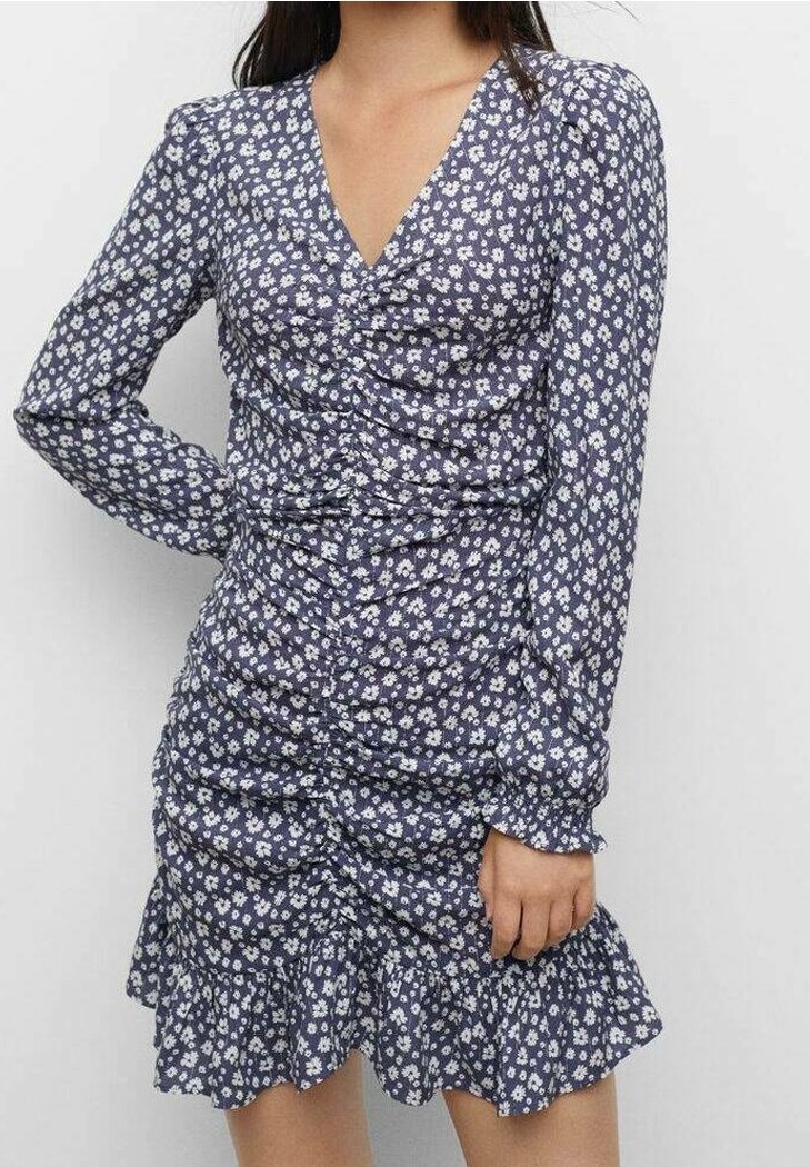 Femme portant une robe manches longues bleu marine avec un motif floral blanc, des détails froncés, un col en V et un ourlet volanté, debout devant un fond uni.