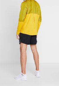 Camiseta de manga larga de Nike en amarillo, con parte superior texturizada y parte inferior lisa. Combinada con pantalones cortos negros y llevada con zapatillas deportivas blancas.