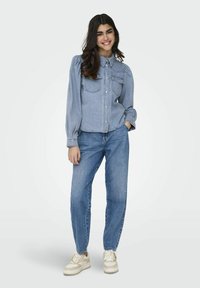 Camicia in denim azzurro chiaro con due tasche frontali, design con bottoni e maniche a sbuffo, abbinata a jeans azzurri chiari e sneakers beige.