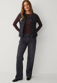 Schwarze gesteppte Weste mit Druckknöpfen, getragen über einem figurnahen lila Langarmshirt. Kombiniert mit lockeren schwarzen Jeans und schwarzen Ballerinas.