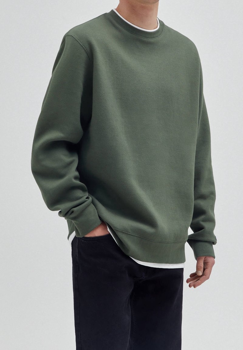 Homme portant un sweat-shirt vert ample à col rond par-dessus une chemise blanche avec un pantalon noir, debout devant un fond gris uni.