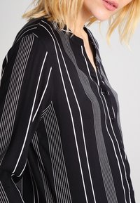 Chemise noire à boutons avec rayures verticales blanches, tissu léger, col et coupe décontractée.
