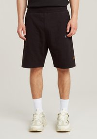 Pantaloni corti neri in cotone con una vestibilità comoda, dotati di tasche laterali, una piccola etichetta logo blu e un dettaglio di cucitura sull'orlo.