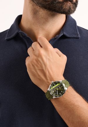 Bärtiger Mann in dunkel strukturiertem Poloshirt, der den Kragen richtet und eine Armbanduhr mit grünem Armband und schwarzem Zifferblatt zeigt.