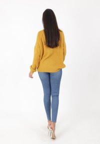 Kobieta z długimi ciemnymi włosami idąca od tyłu, ubrana w żółty sweter w odcieniu musztardowym, niebieskie obcisłe dżinsy i przezroczyste sandały na wysokim obcasie na białym tle.