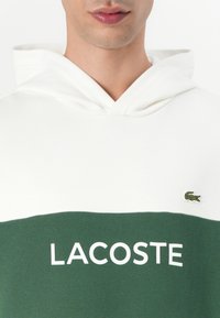 Felpa con cappuccio in misto cotone bianco e verde. Presenta la scritta "LACOSTE" in grande e il logo coccodrillo ricamato sul petto. Tessuto dalla trama liscia.
