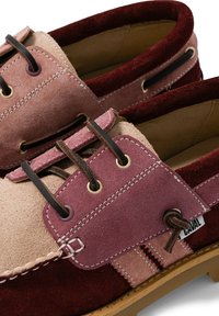 Gros plan sur deux chaussures bateau en daim dans des tons bordeaux, rose et beige avec des lacets noirs et des coutures visibles.