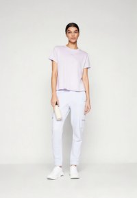 Lavandas krāsas īsroku T-krekls ar vaļīgu piegriezumu, kopā ar gaiši ziliem cargo stila biksēm. Modelis tur baltu ūdens pudeli un valkā baltas sporta kurpes.